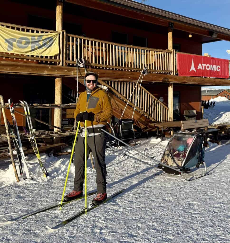 A parent in cross-country ski gear standing with skis and poles next to a kids’ ski trailer outside a lodge