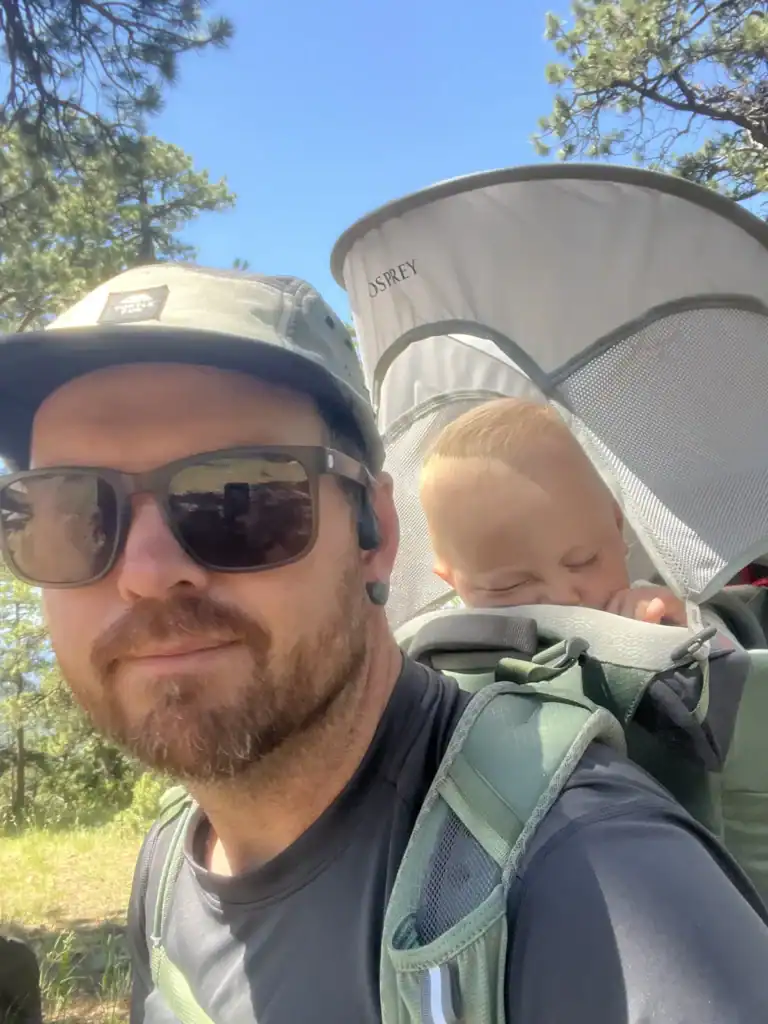 Dad selfie with a toddler asleep under the mesh-sided sunshade of the Osprey Poco Premium child carrier