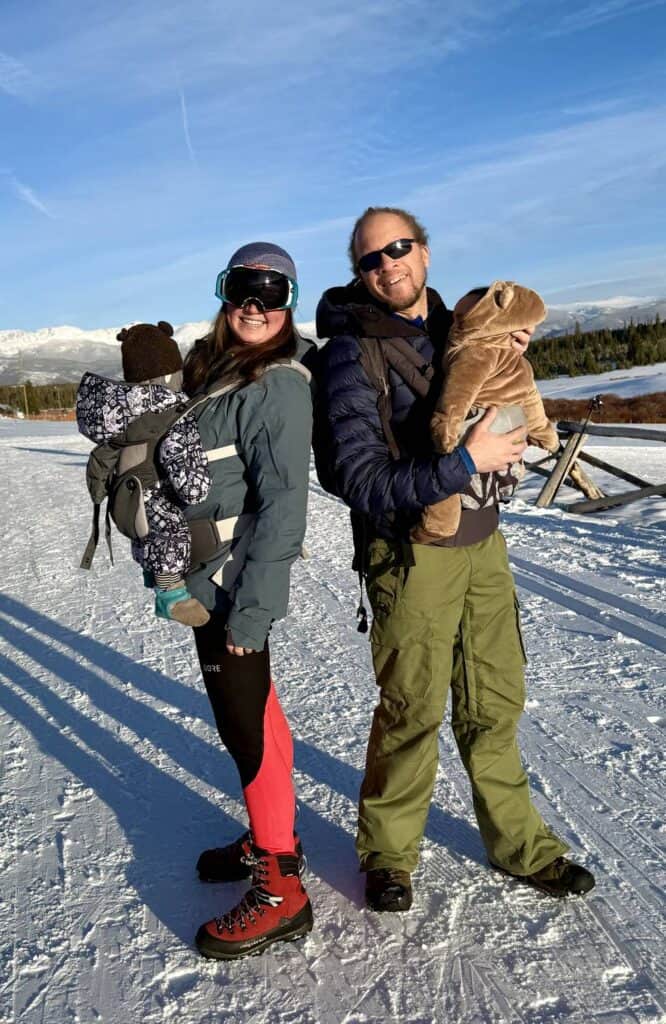 Two parents each carrying a bundled baby in a soft carrier on a snowy field with mountains in the background