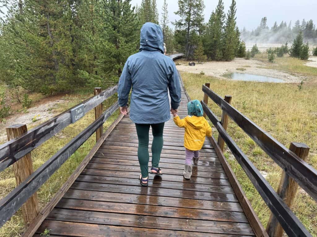 Adult and toddler holding hands on Yellowstone boardwalk, toddler wearing yellow REI Co-op Rainier Rain Jacket