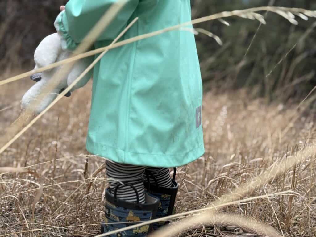 Detail view of lower hem and Reima logo on Lampi Rain Jacket with toddler wearing rain boots