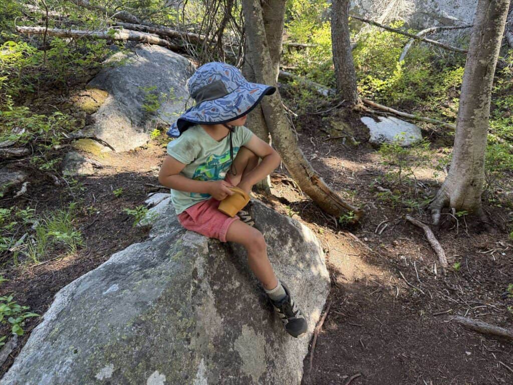 Kid in a sun hat sitting on a granite boulder in the forest wearing Salomon Speedcross kids shoes