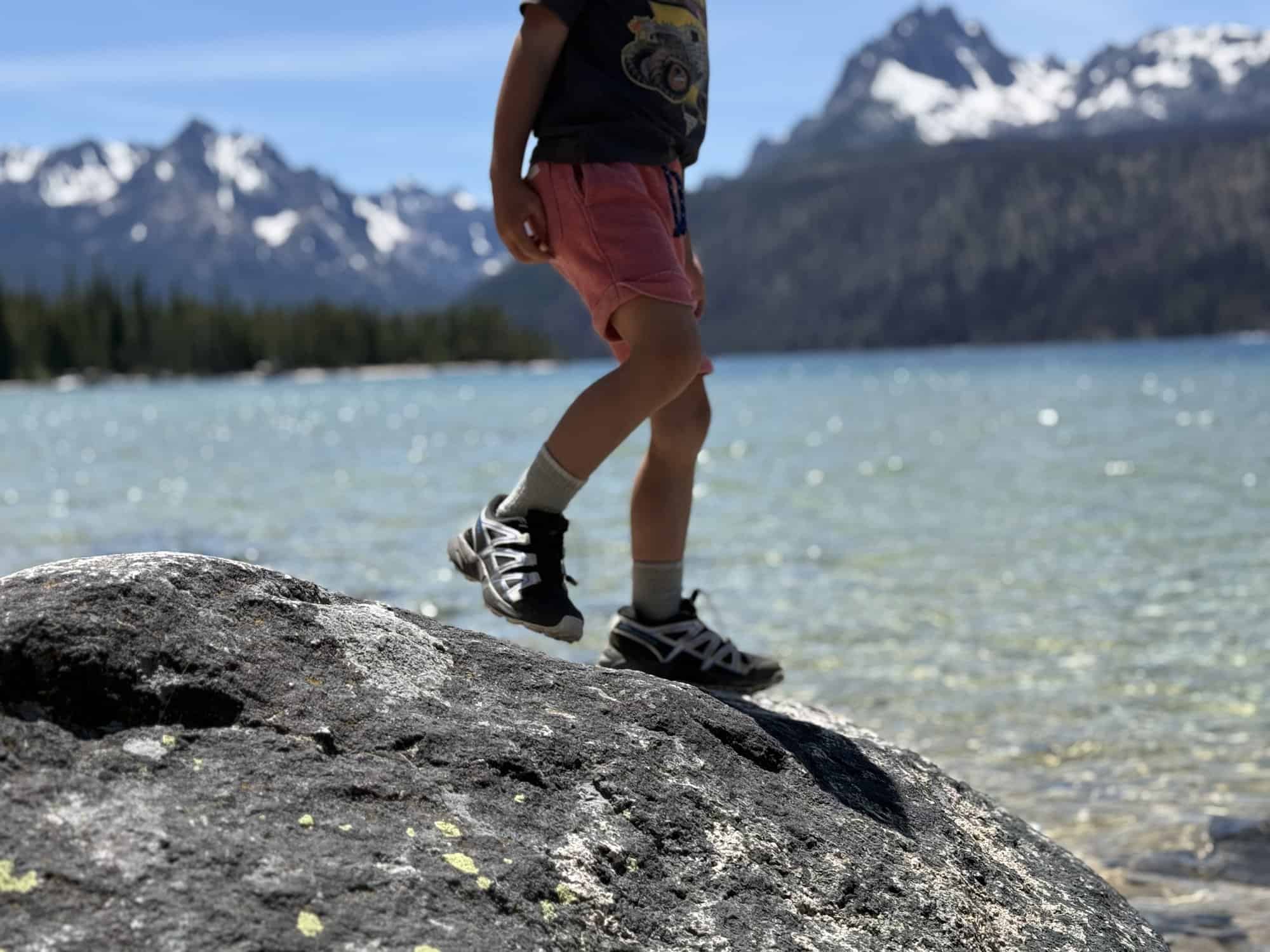 Salomon Speedcross kids shoes stepping across a granite boulder at Redfish Lake, Idaho with the Sawtooth Mountains behind