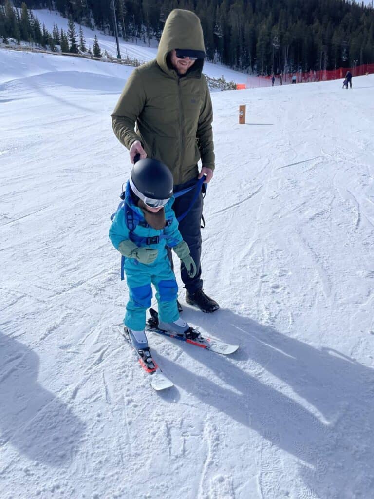 A toddler in a full snowsuit, helmet, and goggles learning to ski with a harness strap held by a parent