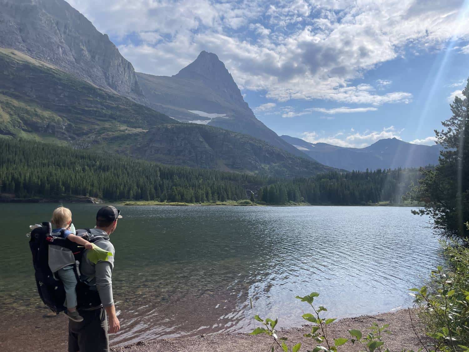 A father carrying a toddler in a backpack hiking carrier beside a mountain lake, with dramatic peaks in the background