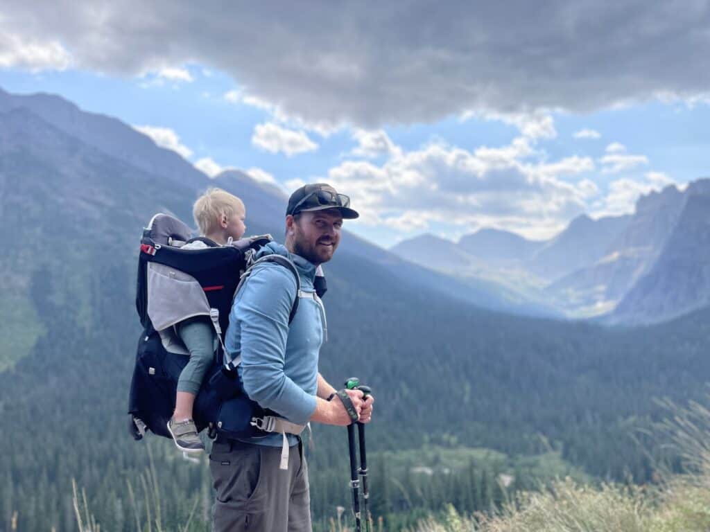 A father carrying a toddler in a backpack hiking carrier, using trekking poles, with dramatic mountain cliffs in the background