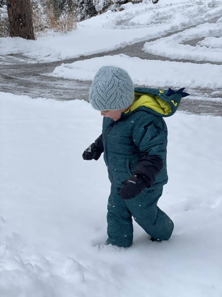 A toddler walking through deep snow in a full snowsuit, knit beanie, and mittens
